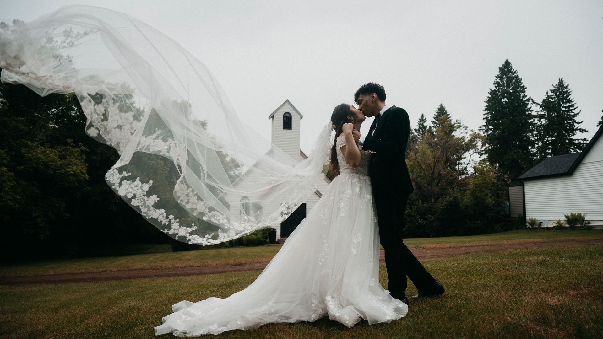 A bride and groom are kissing in front of a church with their veil blowing in the wind.