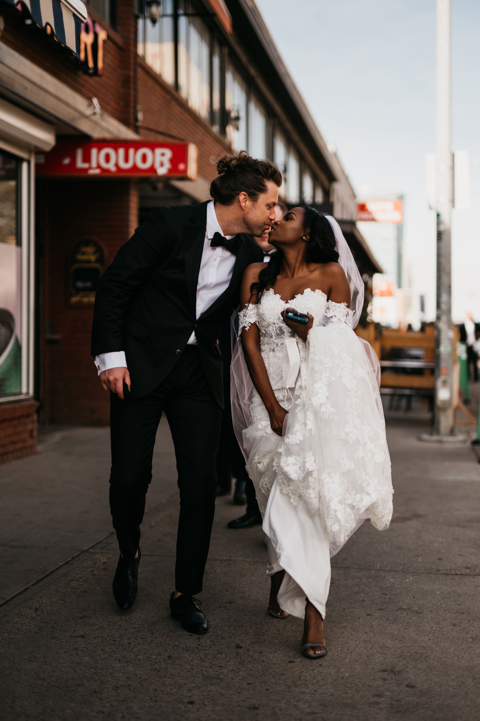 A bride and groom are kissing on the sidewalk in front of a liquor store.