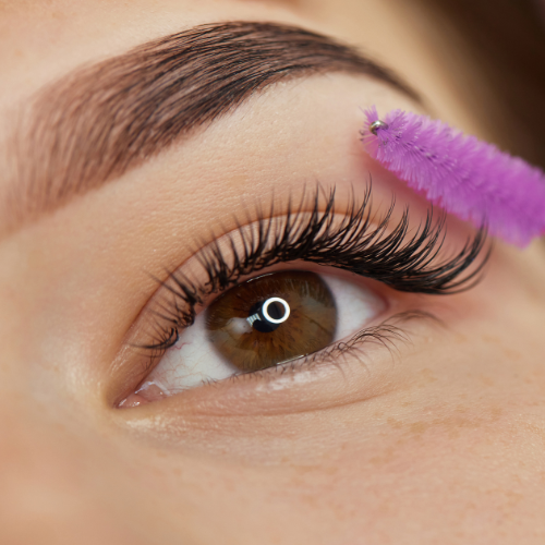 Close-up of eye with long, dark lashes being brushed with a purple mascara wand. Light brown eyebrow.