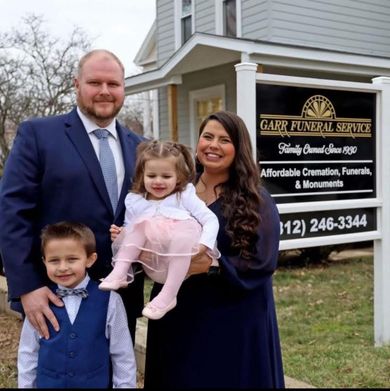 Family posing in front of Garr Funeral Service sign; woman holds child, man stands with another child.
