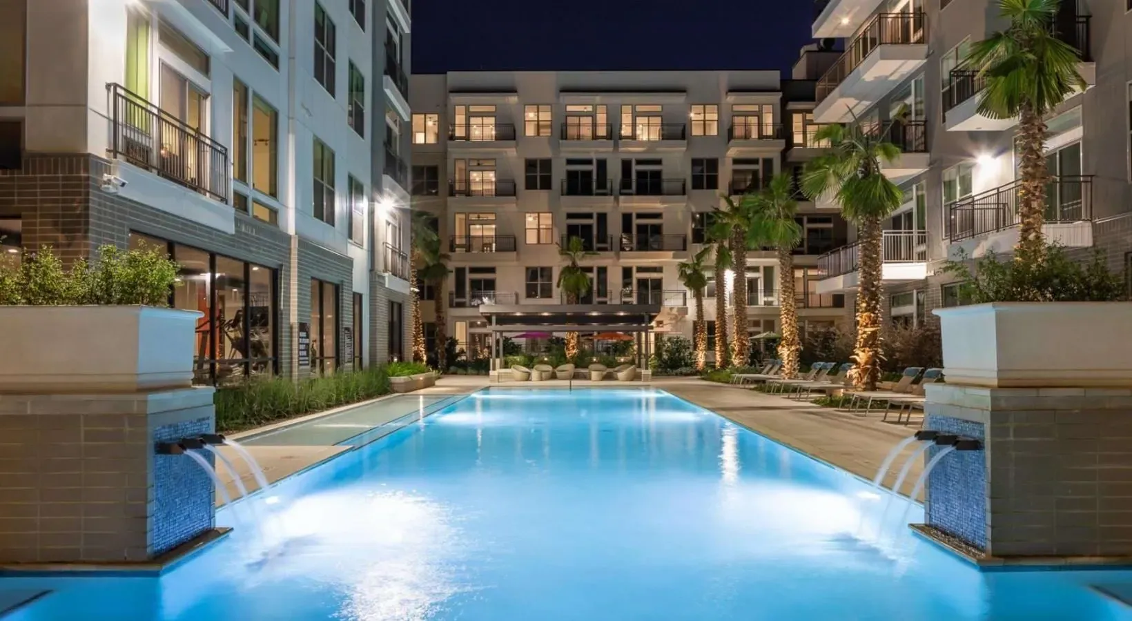 Luxury apartment complex courtyard with an illuminated swimming pool at night, palm trees, and seating areas.