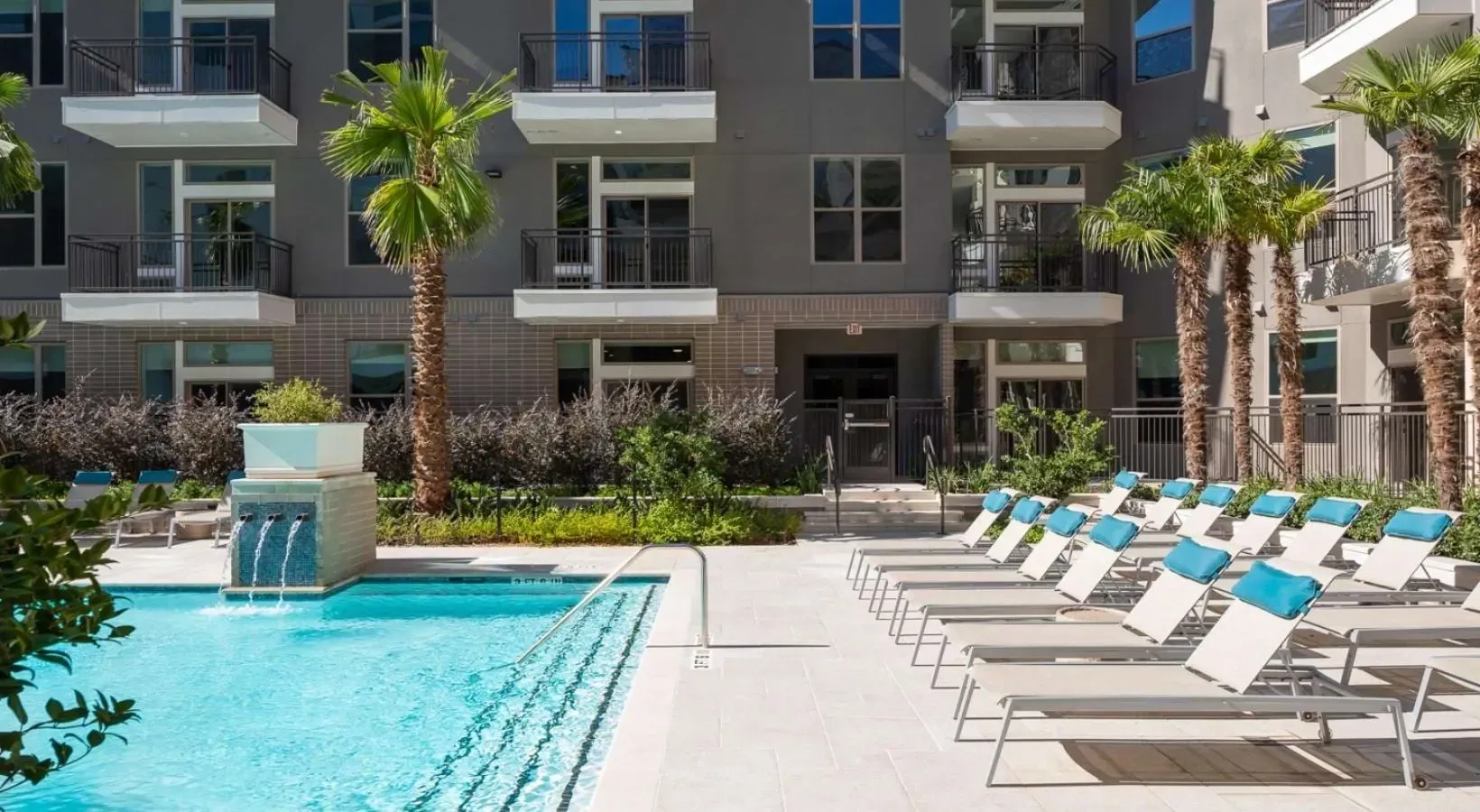 Resort-style outdoor swimming pool with lounge chairs and palm trees next to a modern apartment building.