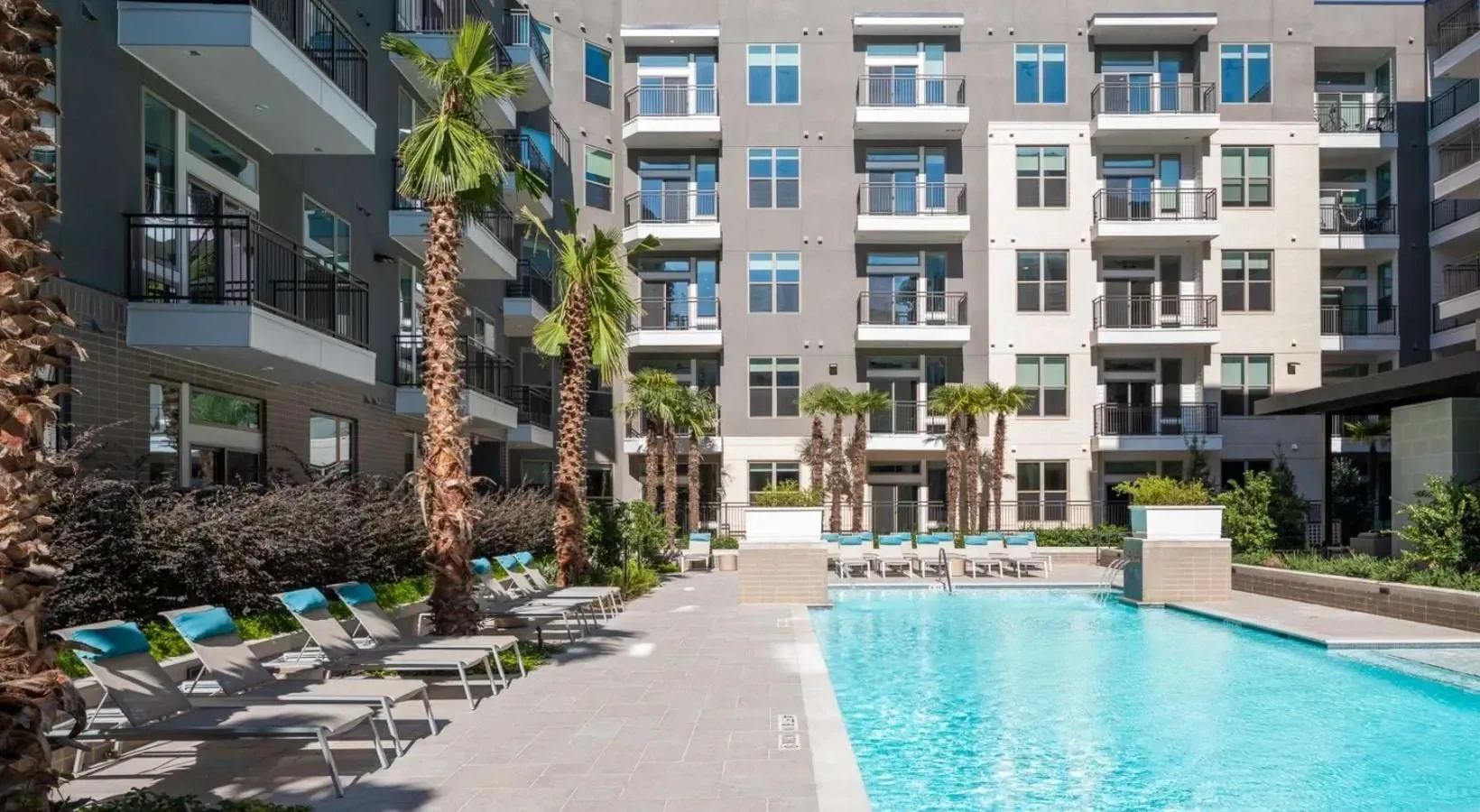 Resort-style outdoor swimming pool with lounge chairs and palm trees next to a modern apartment building.