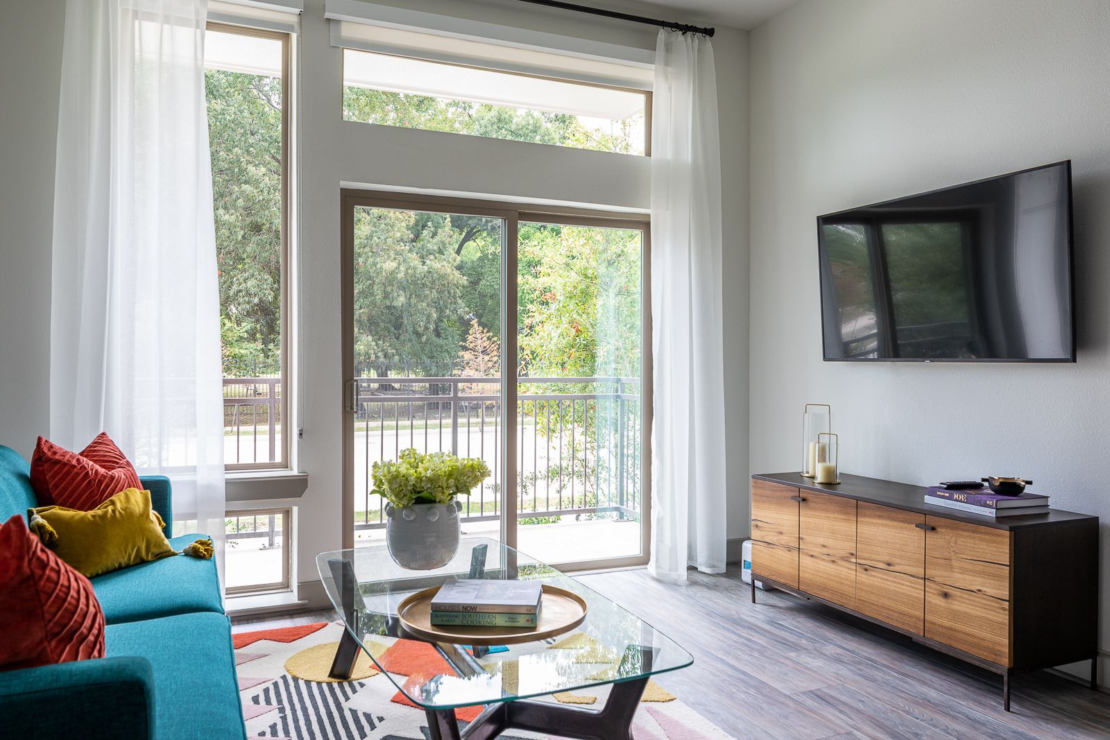 Bright living room with teal sofa, glass coffee table, TV, and balcony access with a scenic view.