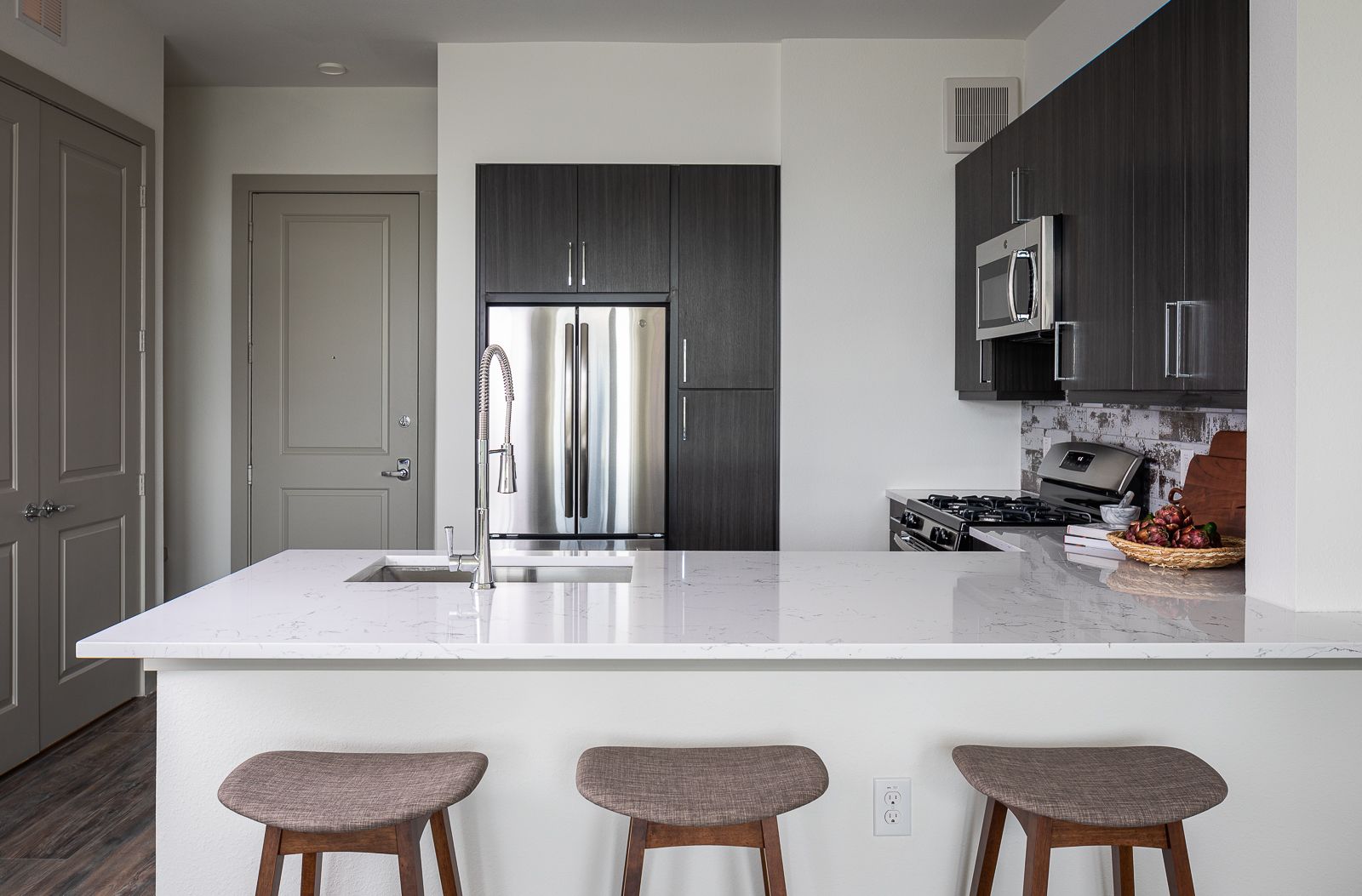 Modern kitchen island with three stools and quartz countertop, stainless steel appliances, and dark wood cabinets.