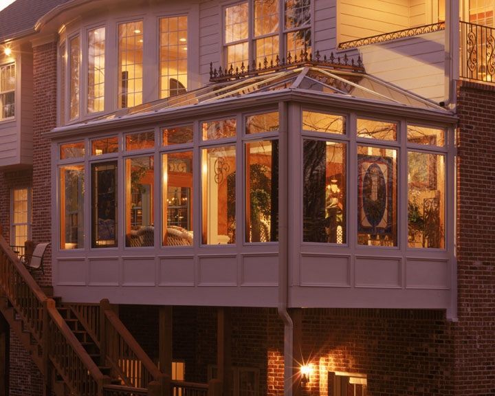 Sunroom addition on a house; glass windows and roof, exterior brick wall, wooden deck and stairs.