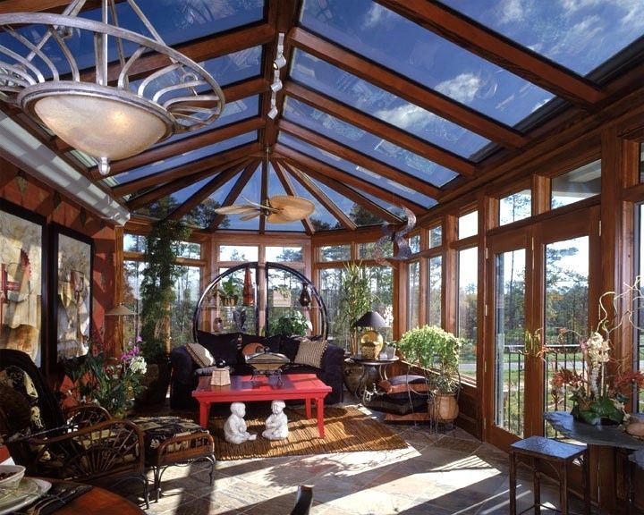 Sunroom with wood frame, glass roof, furnished with sofa, plants, and table, overlooking a yard.