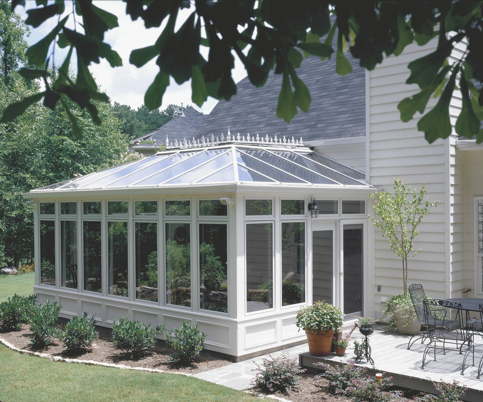 Sunroom attached to a house with glass windows and roof. Plants and patio furniture are visible.