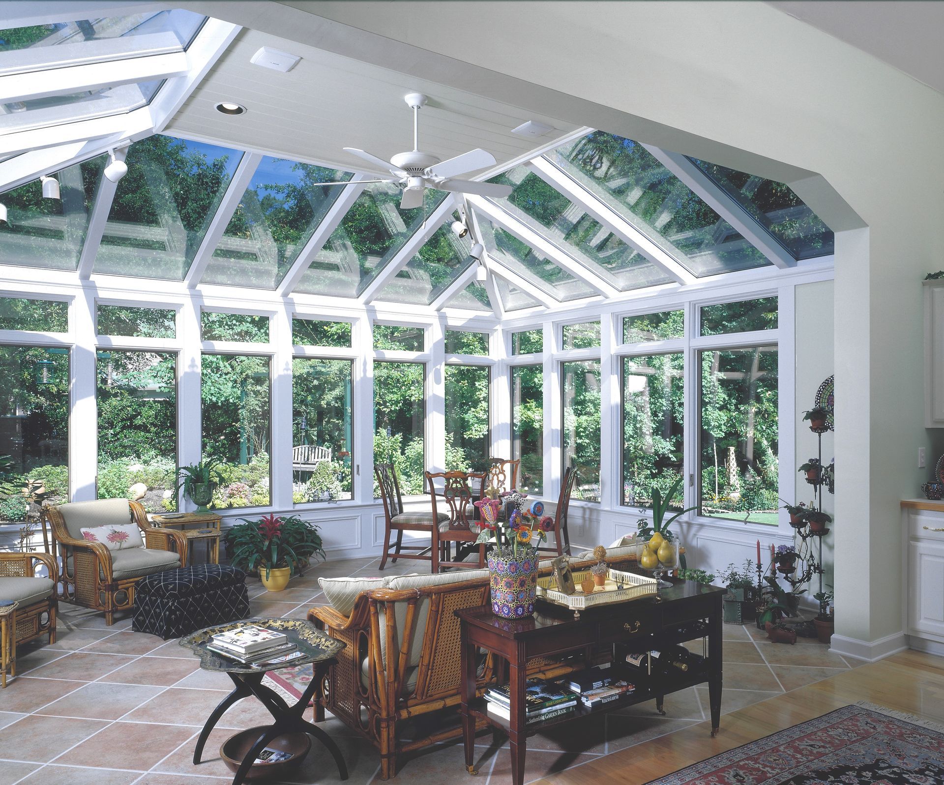 Sunroom with glass roof and walls, furnished with seating and a dining table, overlooking a lush garden.