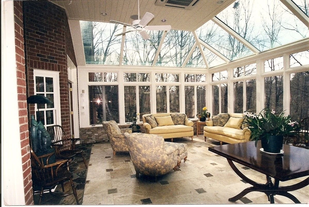 Sunroom with glass ceiling and walls, furnished with sofas, chairs, and a table. Brick wall on the left.