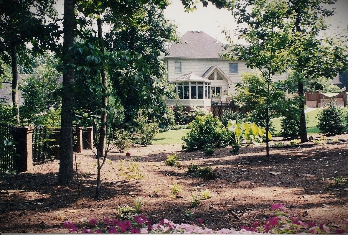 Large house with light siding and dark roof, seen through trees and a garden bed.
