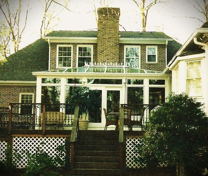 House with sunroom, deck, and brick chimney. Green roof, white trim, trees in the background.