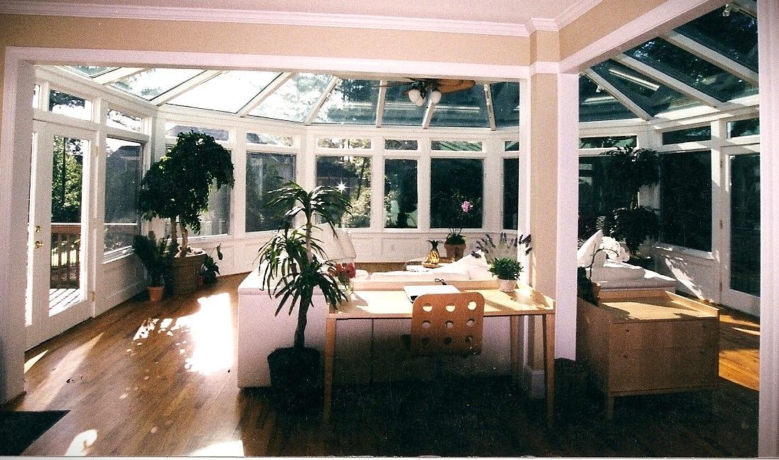 Sunroom with glass walls and ceiling, hardwood floors, and potted plants. Furnished with a desk, cabinets, and a chair.