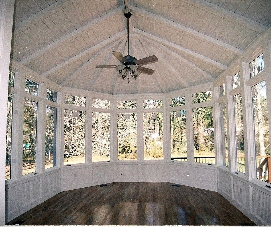 Sunroom with white paneling, wooden floor, large windows, and a ceiling fan, overlooking a wooded view.