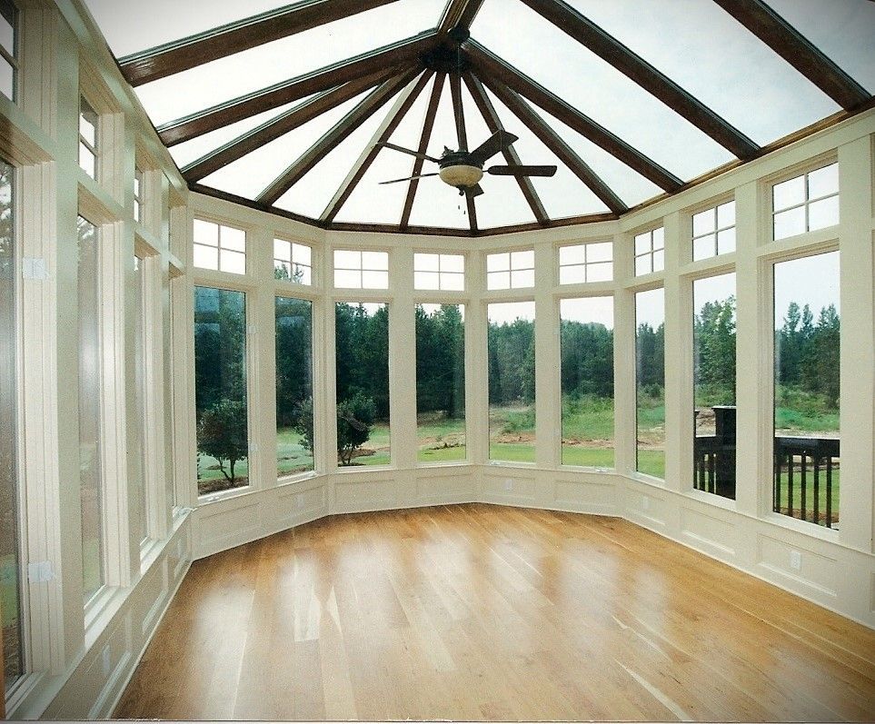 Sunroom with angled glass roof, multiple windows, wooden floor, and ceiling fan, overlooking a green landscape.