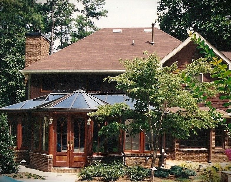 Sunroom addition with glass roof and wood trim on a brick house, surrounded by trees.