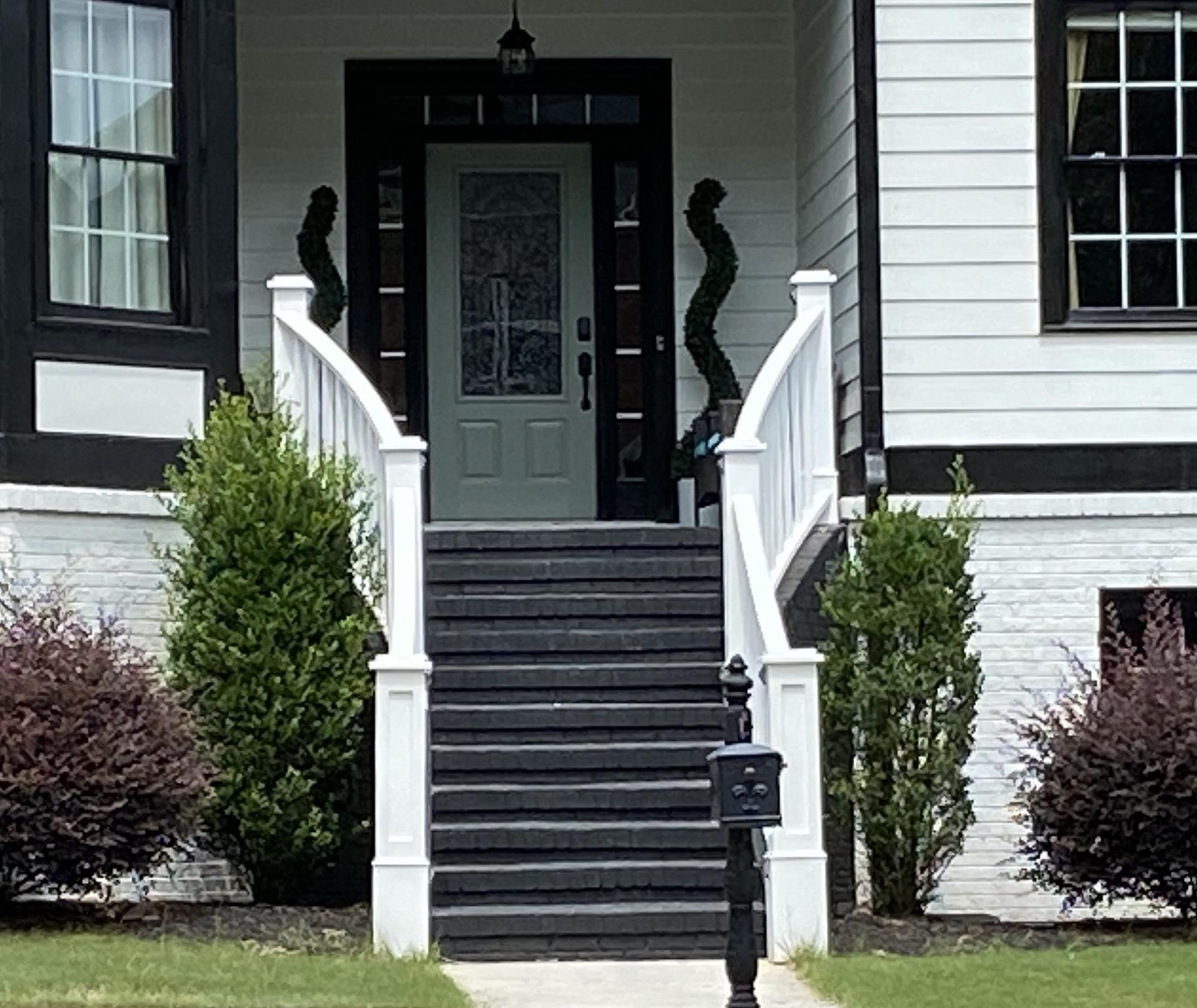 Black and white house entrance with steps, white railing, and decorative shrubs.