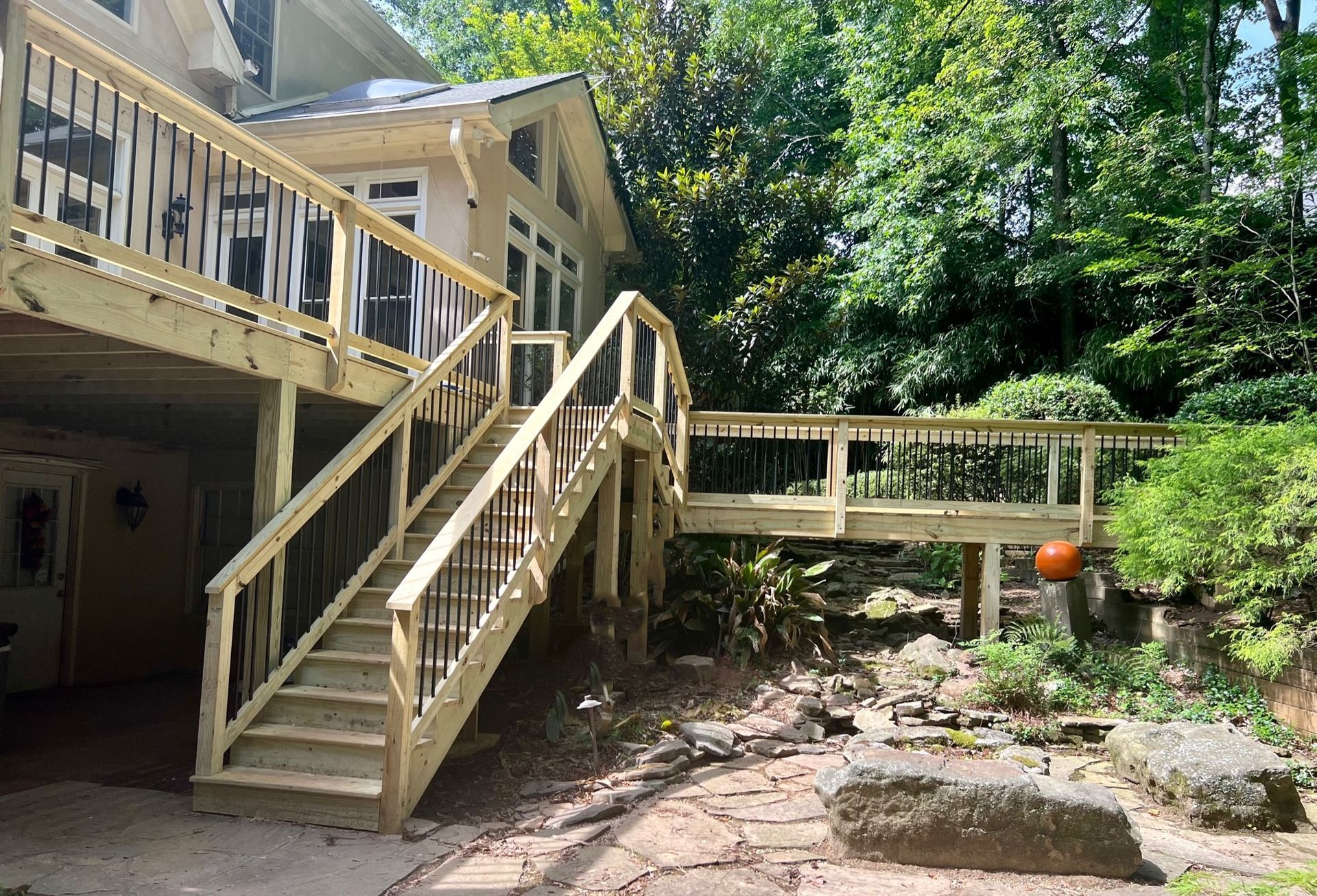 Wooden deck with stairs leading to a house. Black railings, landscaping, and trees are visible.
