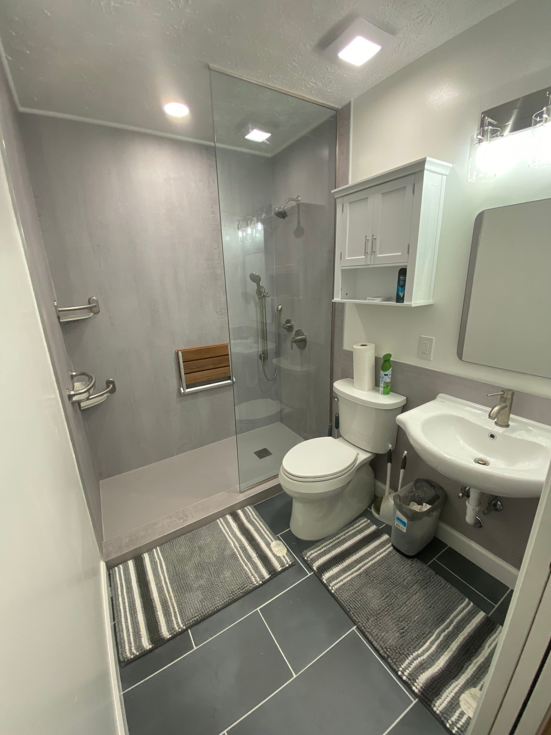 Modern bathroom with gray walls, glass shower, white toilet, and sink. Two gray rugs on the floor.