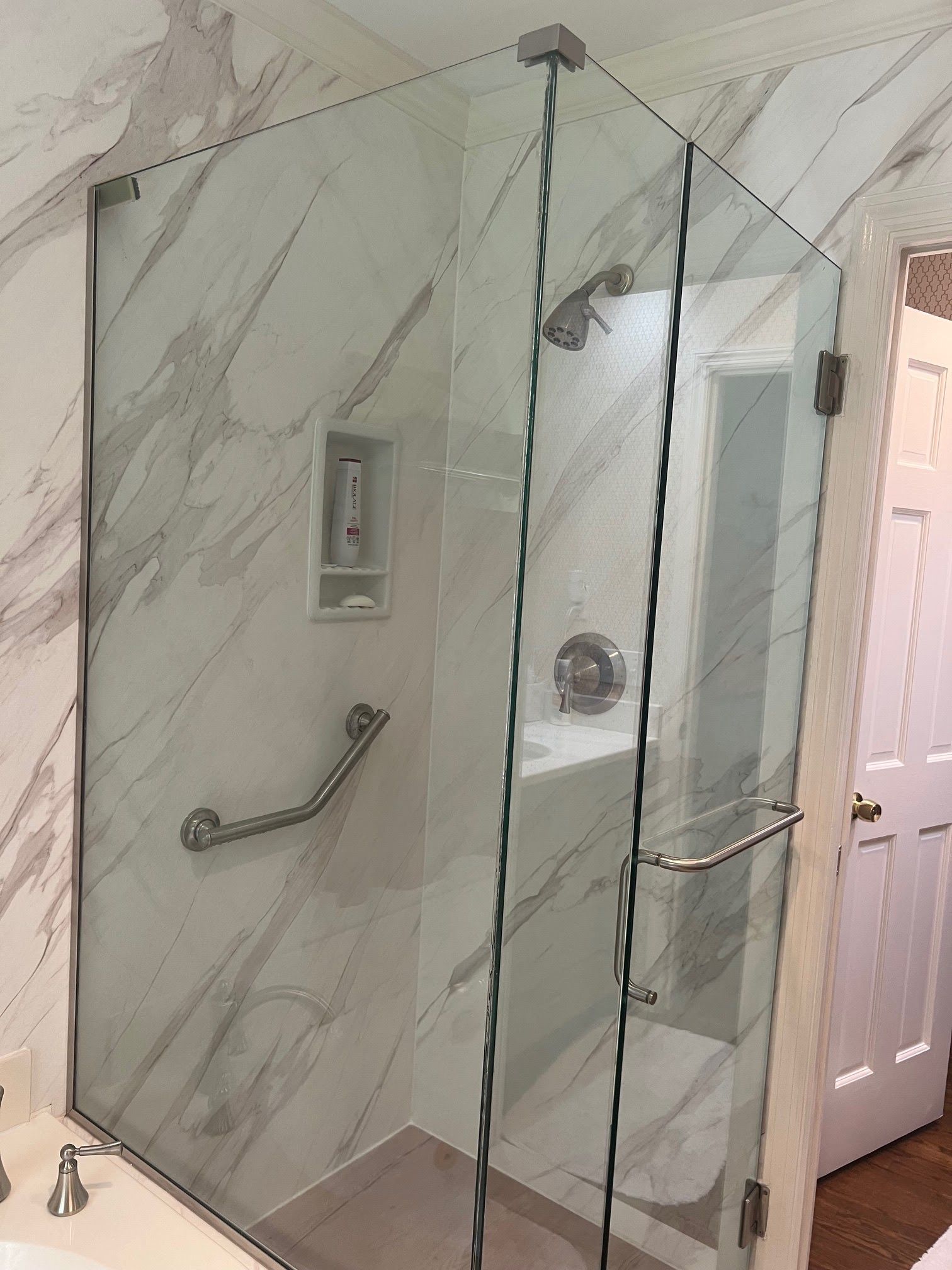 Glass shower enclosure in a bathroom with white marble tile and grab bar.