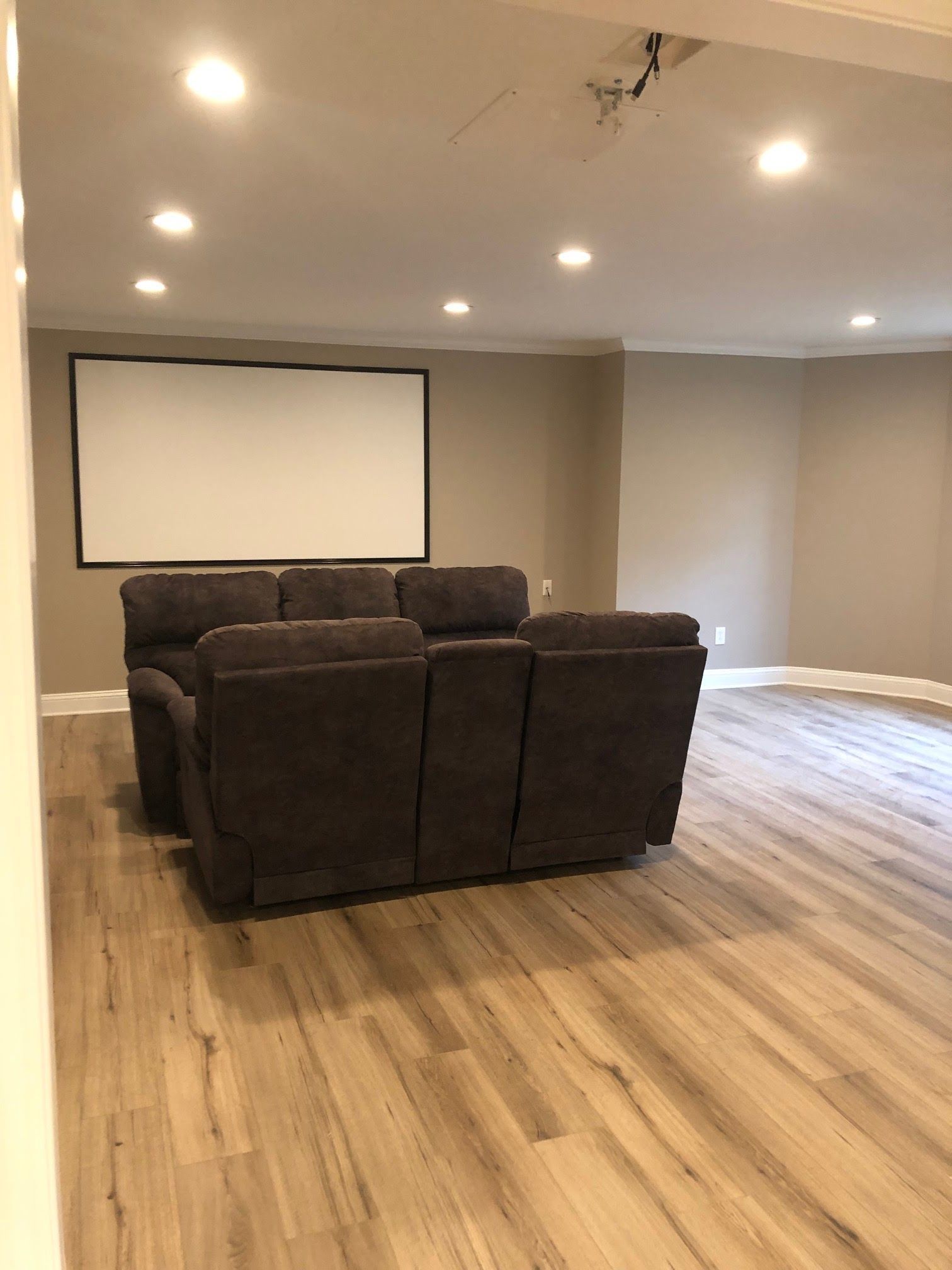 A dark brown reclining couch faces a projection screen in a room with light wood floors and recessed lighting.