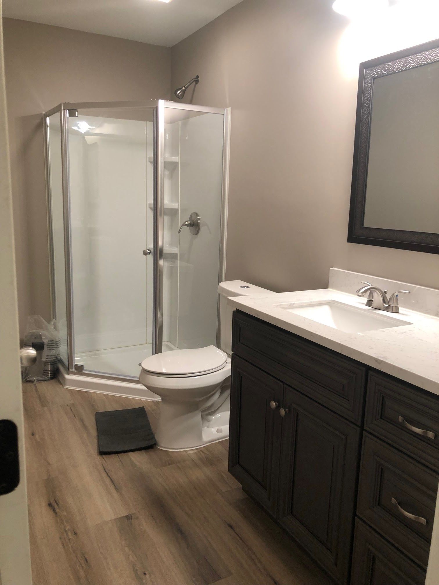 Bathroom with a shower, toilet, vanity, and wood-look flooring. Neutral walls, dark brown cabinets, and a black framed mirror.