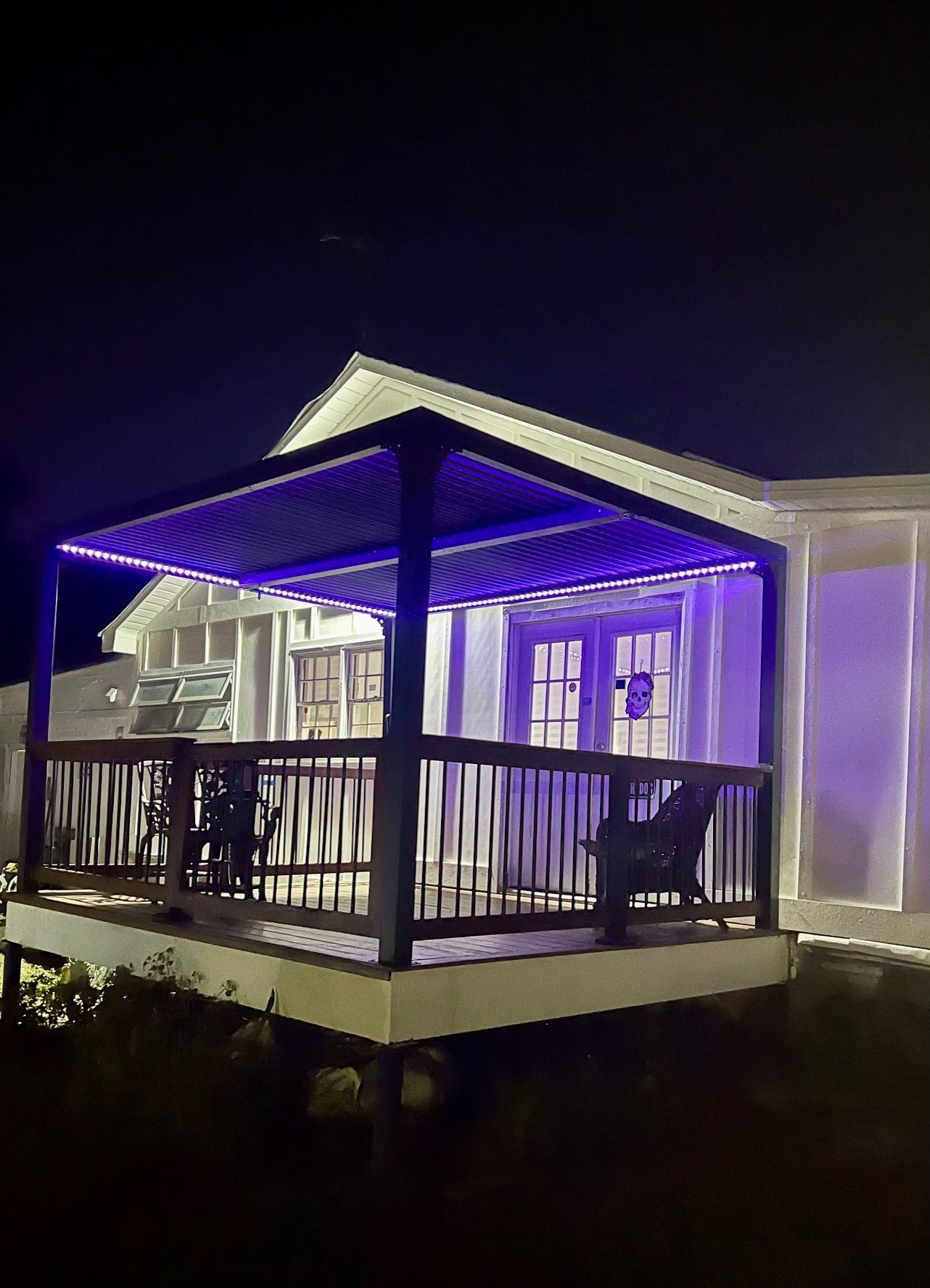 A patio with purple LED lights on the pergola at night. Dark wood deck and railing, white house in background.