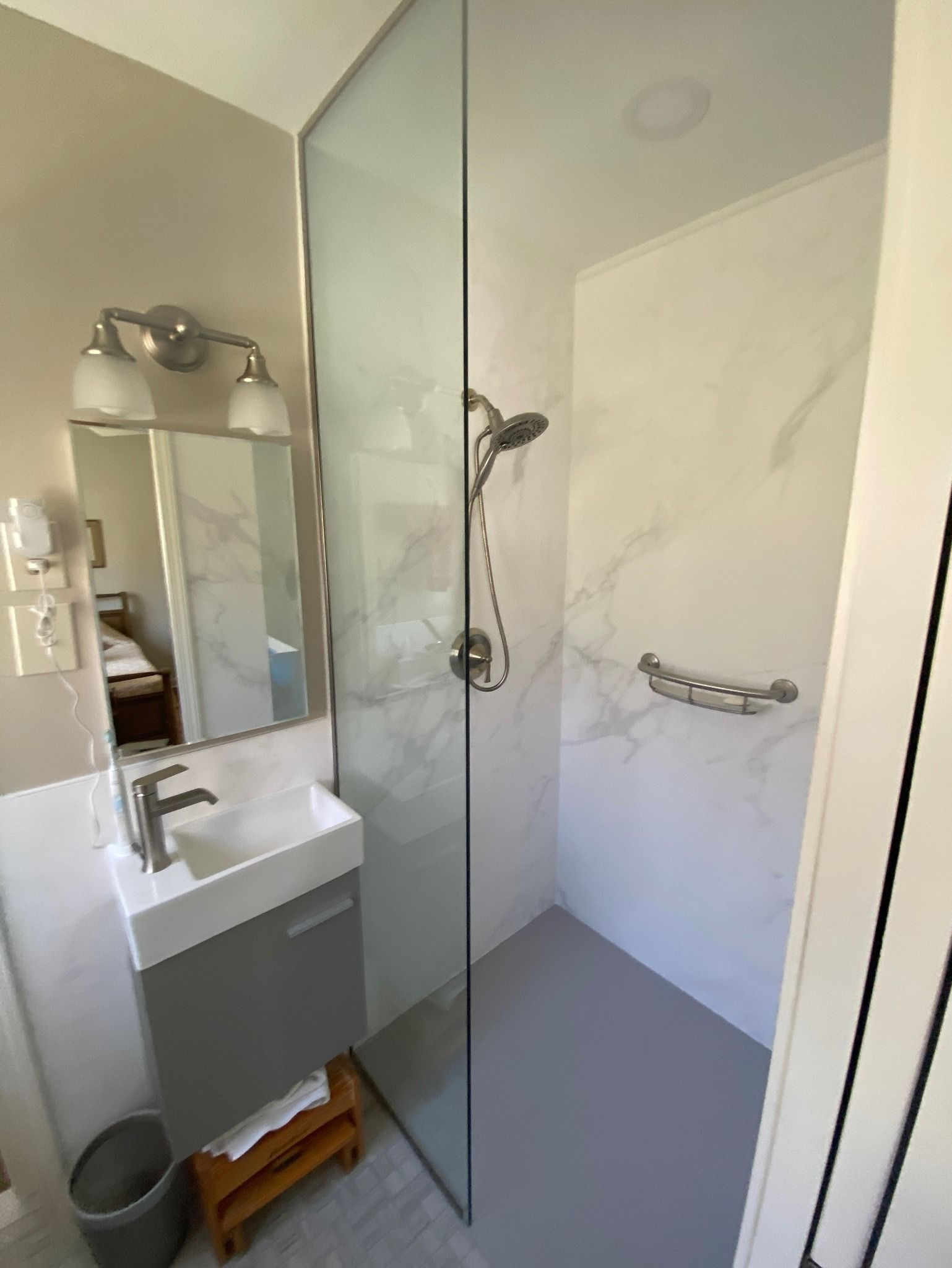 Small bathroom with walk-in shower. Gray vanity with sink, mirror, and a small wooden stool. Shower has grab bar.
