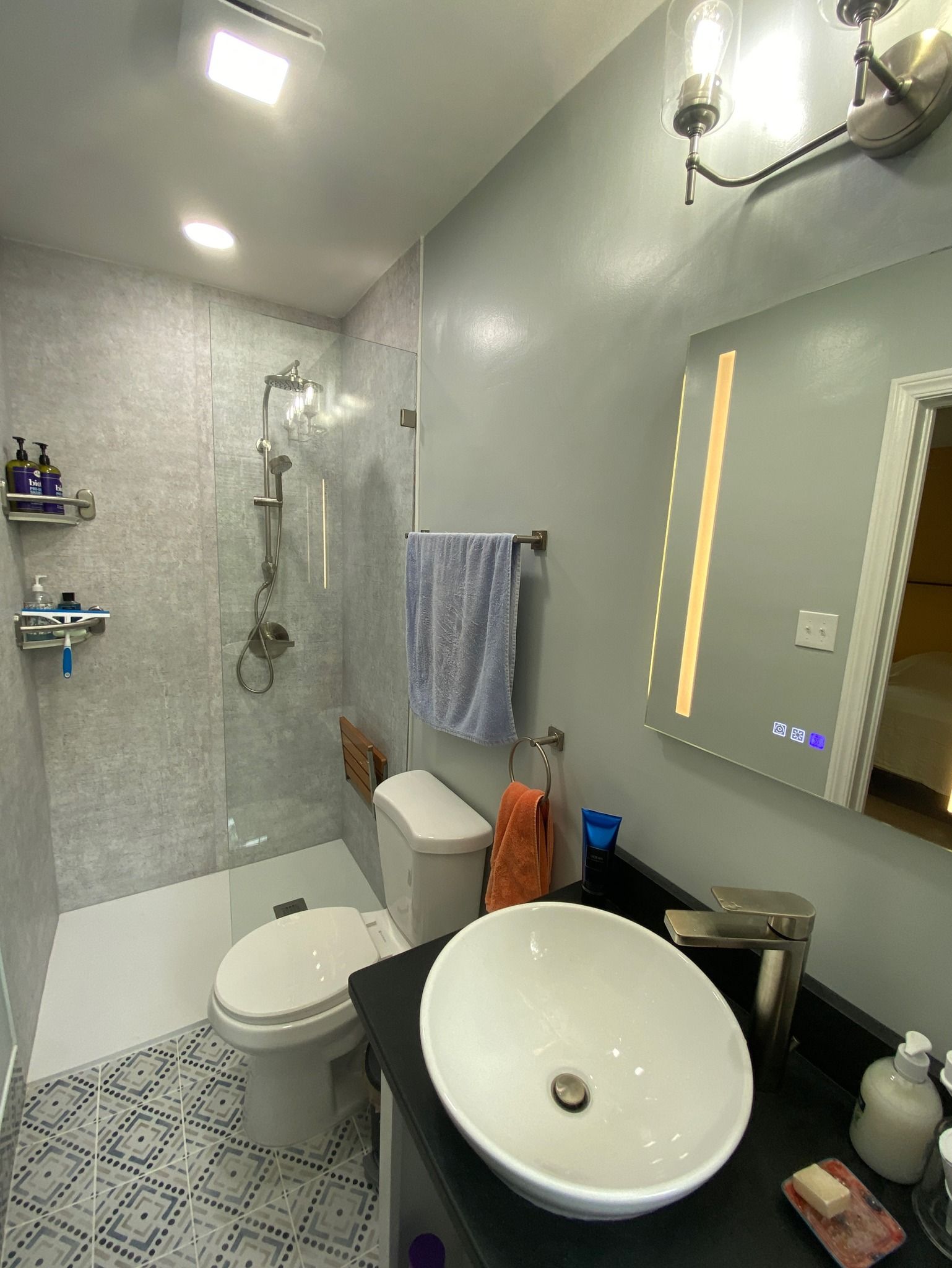Modern bathroom with shower, toilet, and vessel sink. Gray walls, patterned floor tiles, and a lit mirror.