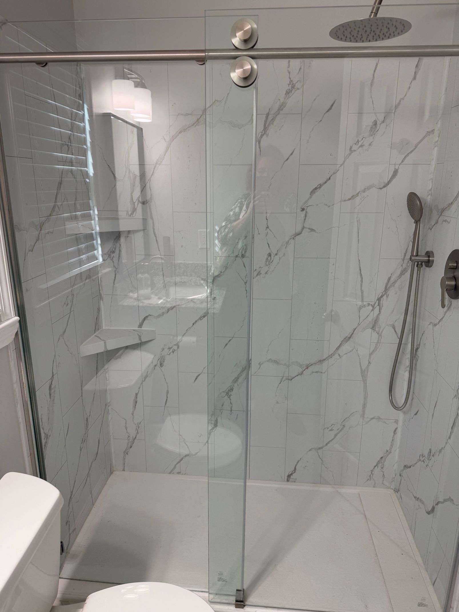 Glass shower enclosure with white marble-like tile walls. Chrome hardware and showerhead are visible.