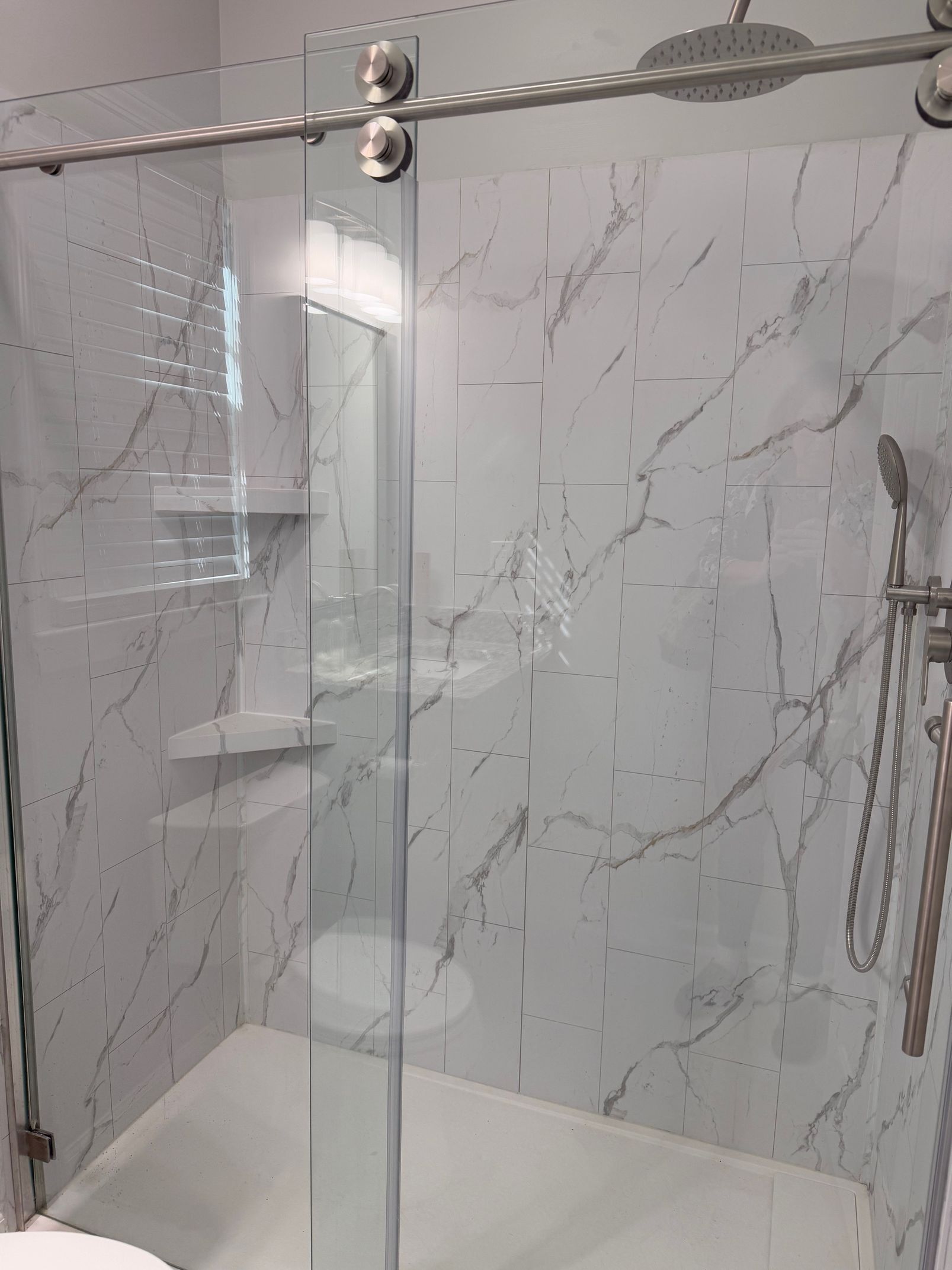 Glass shower with white marble-patterned walls. Silver fixtures, sliding door, built-in shelves.