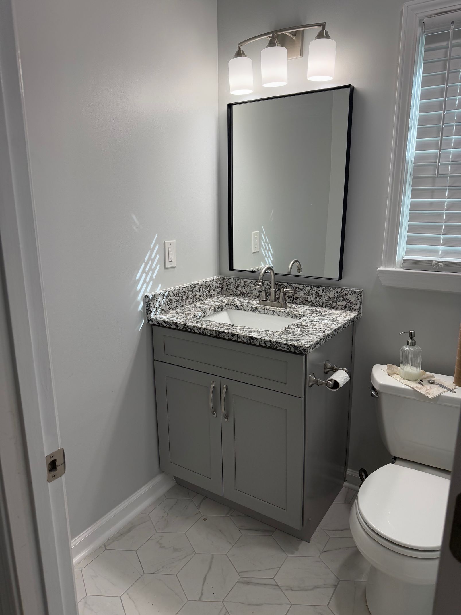 Bathroom with gray vanity, granite countertop, mirror, toilet, and window with blinds.