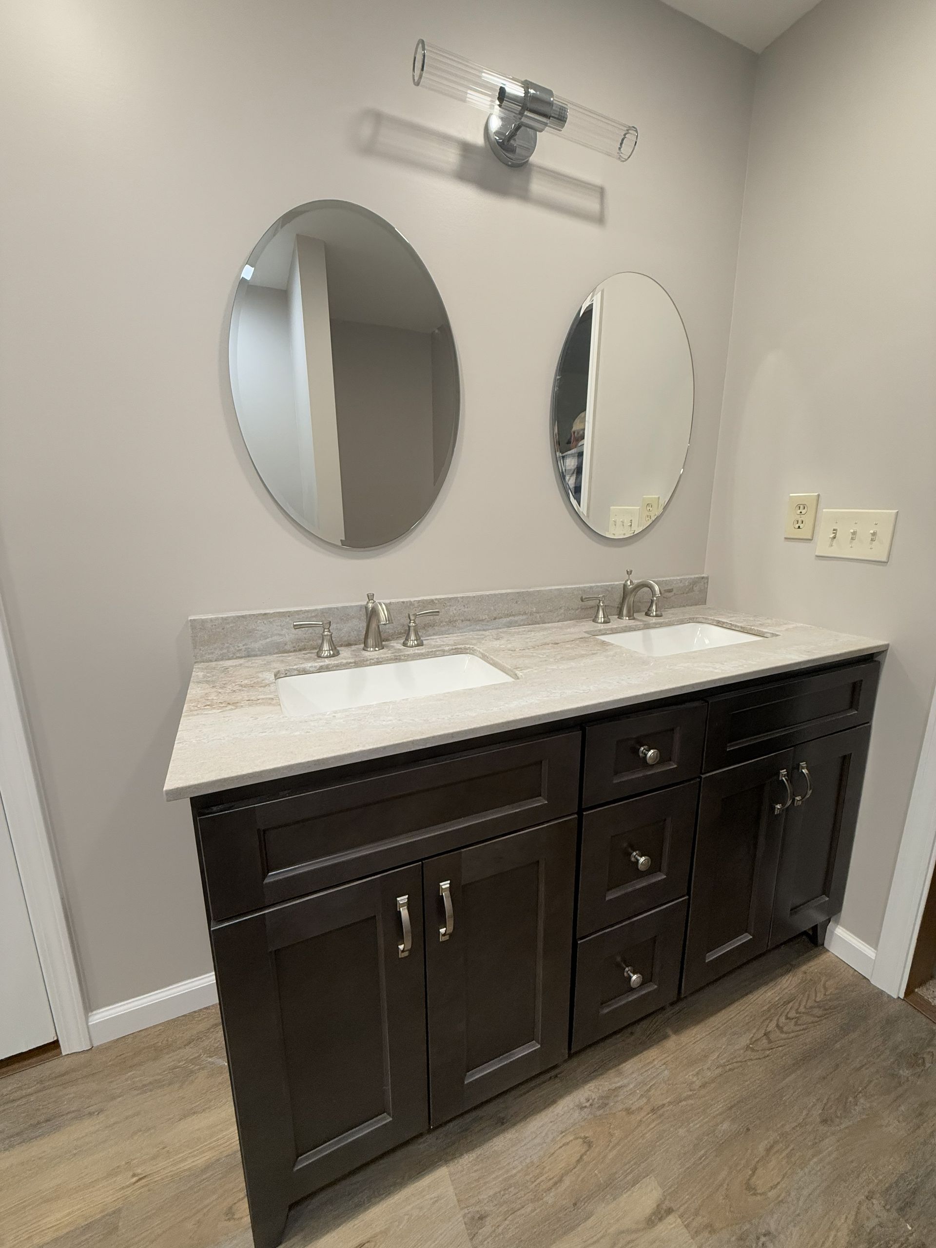 Bathroom vanity with dark brown cabinets, two oval mirrors, and light gray walls.