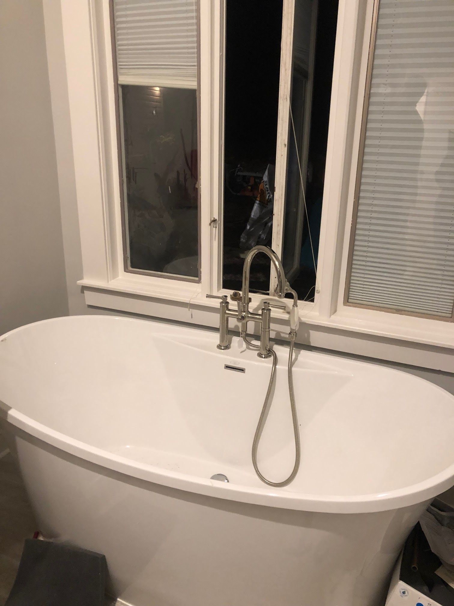 White freestanding bathtub with silver faucet, under a window with blinds.