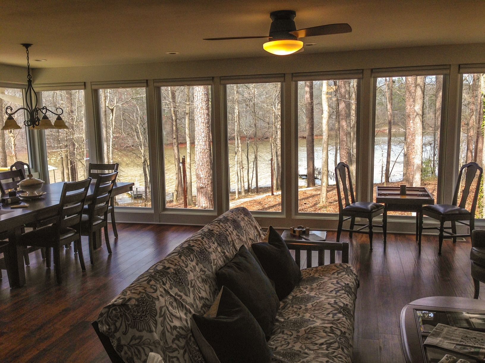 Living room with large windows overlooking trees and water; dark wood furniture; sofa, dining table, chairs.