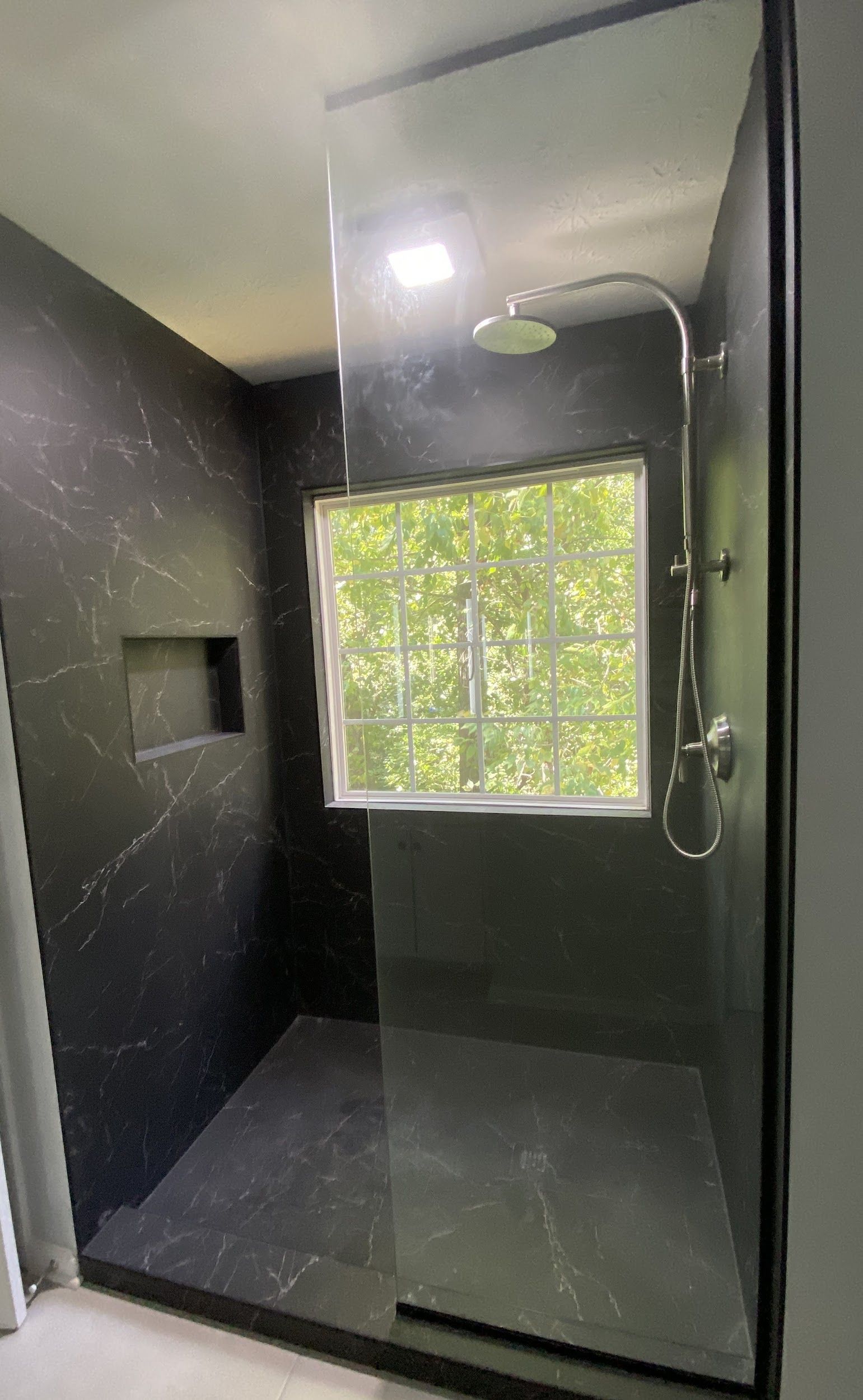 Dark tiled shower with glass door and rain showerhead, window in the back.