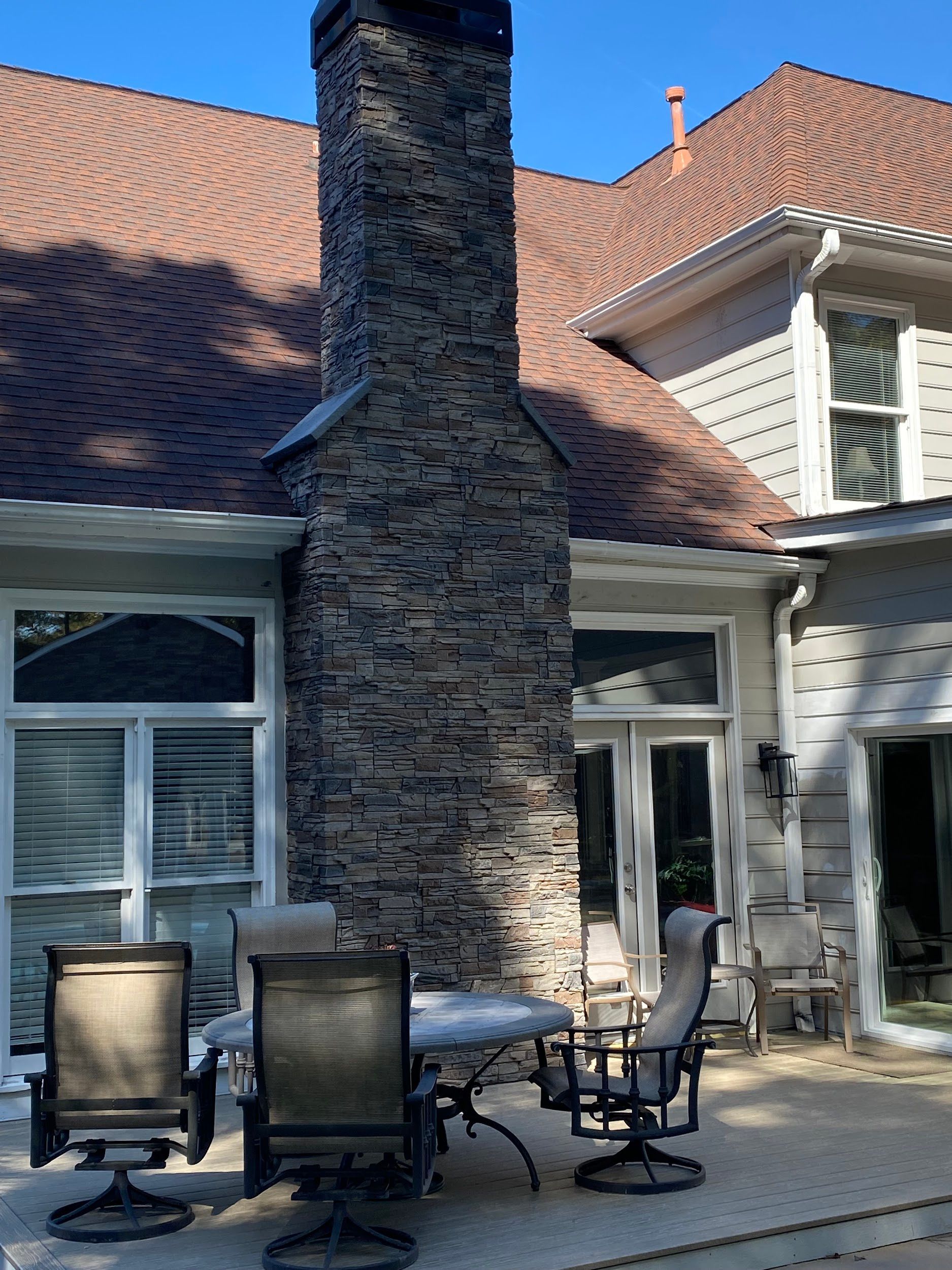 Stone chimney on a house with a brown shingle roof, patio with chairs and table on a sunny day.