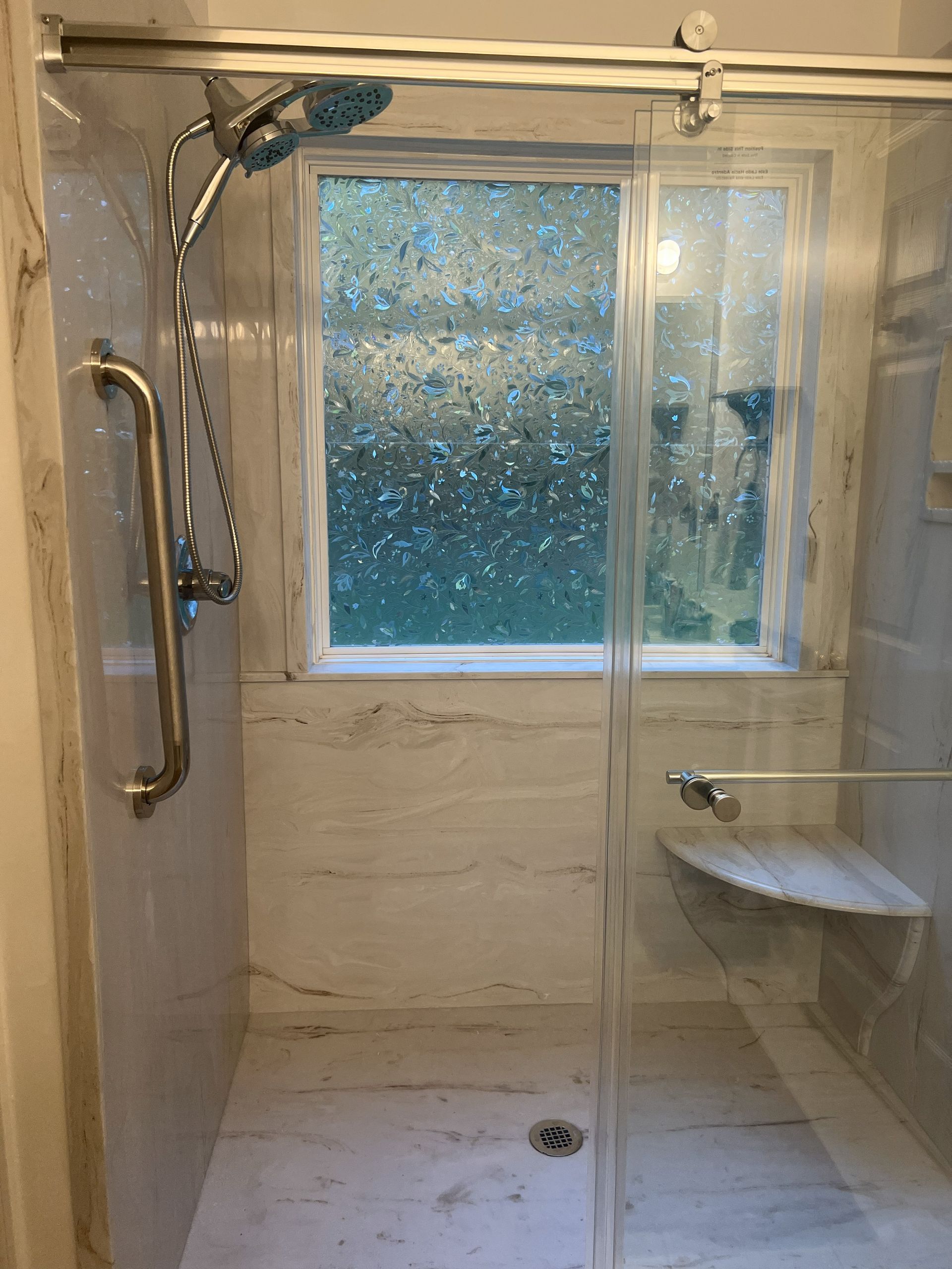 Shower with glass door, marble-like tiles, a window, grab bar, and built-in bench.