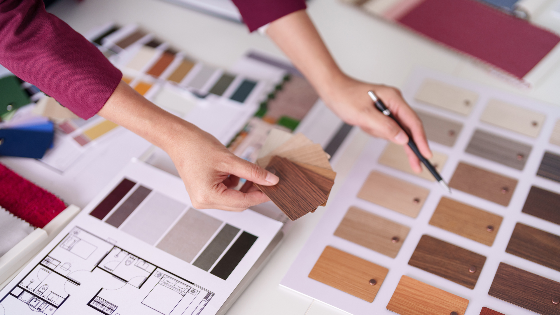Person's hands selecting wood swatches, holding a pen over a color palette and floor plan on a desk.