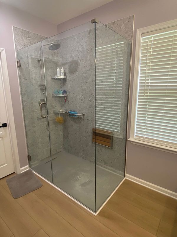 Modern glass shower with gray tiled walls, stainless steel fixtures, and wood bench near window.