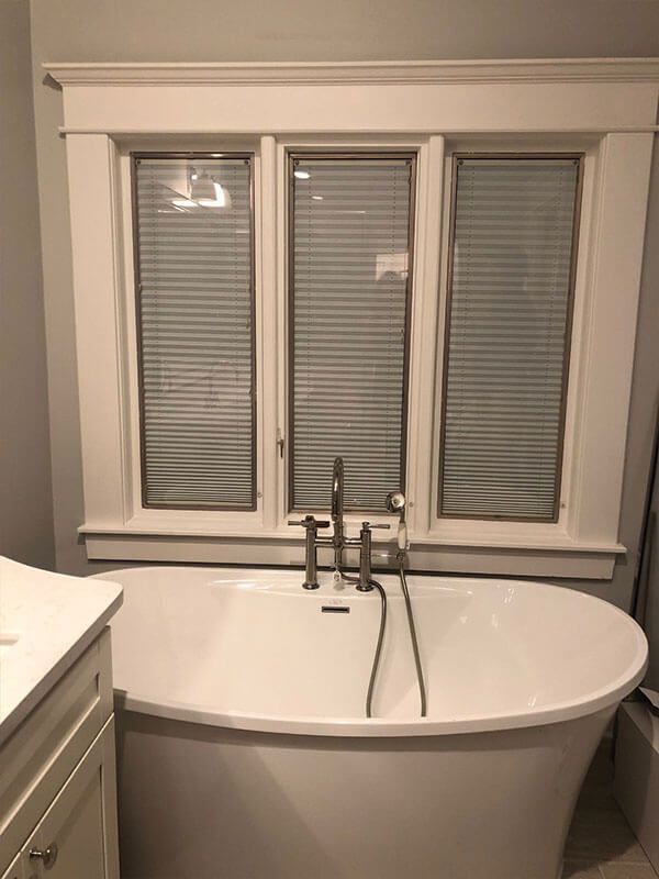 White bathtub beneath a window with closed blinds, in a bathroom with light gray walls and white trim.