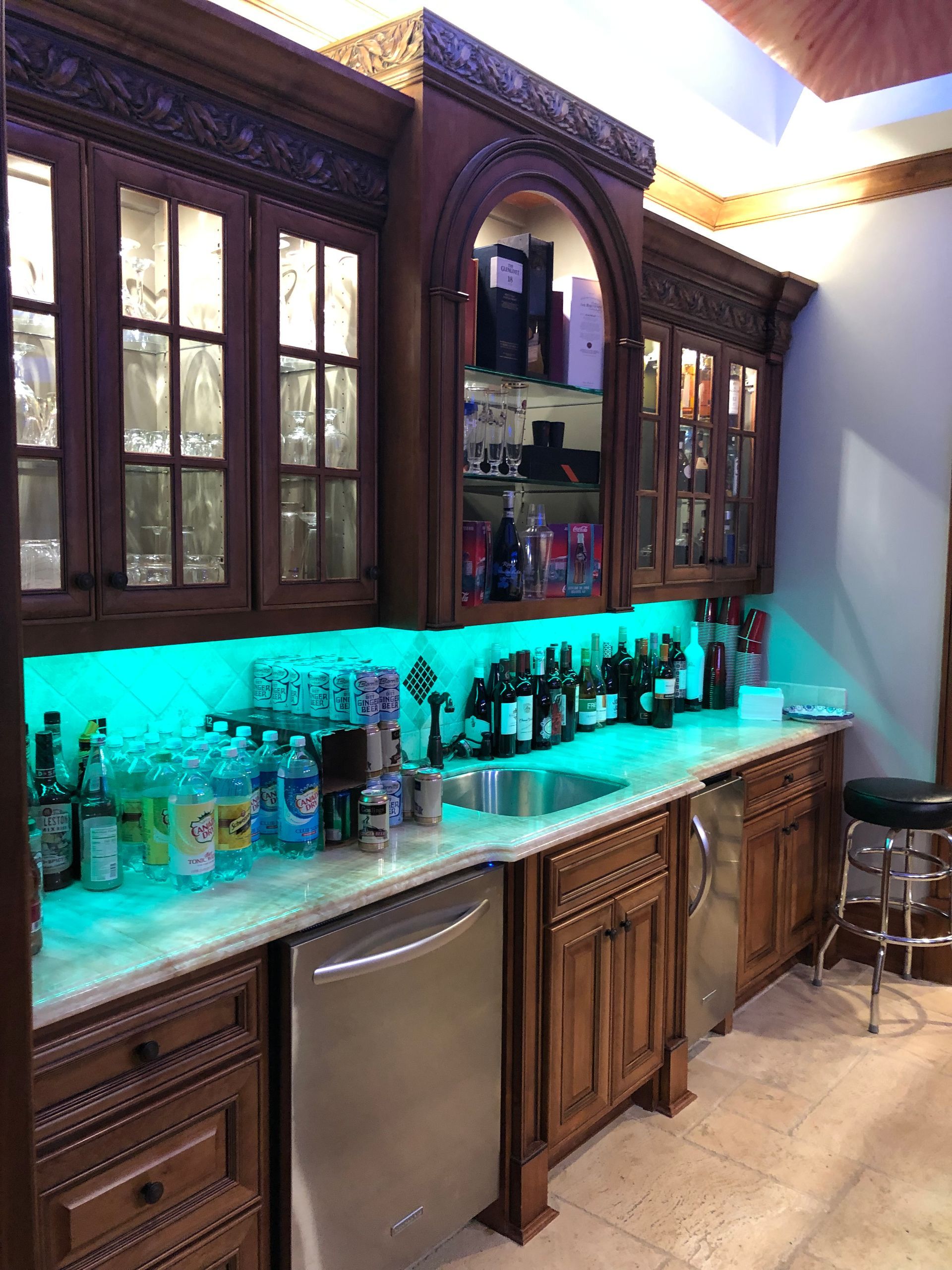 Wooden home bar with glass-front cabinets, bottles, a sink, and an under-counter dishwasher. Teal lighting illuminates the countertop.