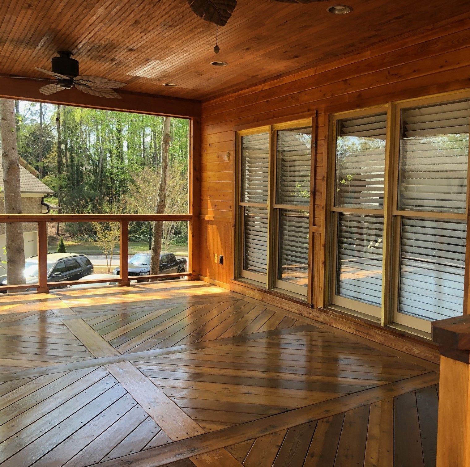 Wooden screened porch with windows, a ceiling fan, and herringbone floor, overlooking trees and a driveway.