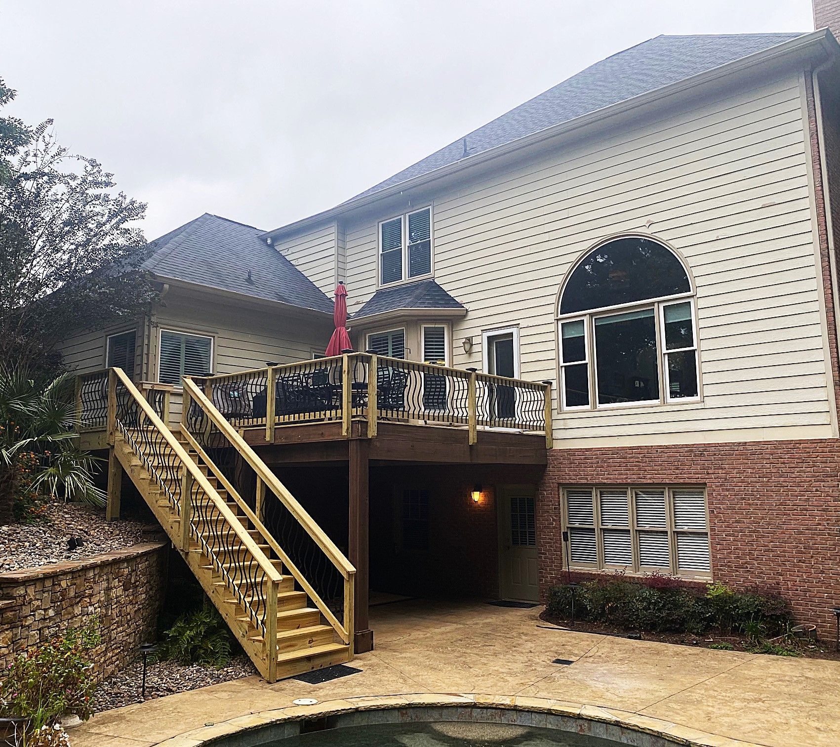 Back of a two-story beige house with a wooden deck and staircase. A pool is in the foreground.