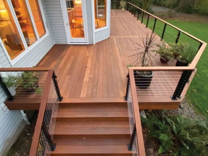 Wooden deck with cable railing and stairs leading to a house, potted plants on the deck, and green lawn.