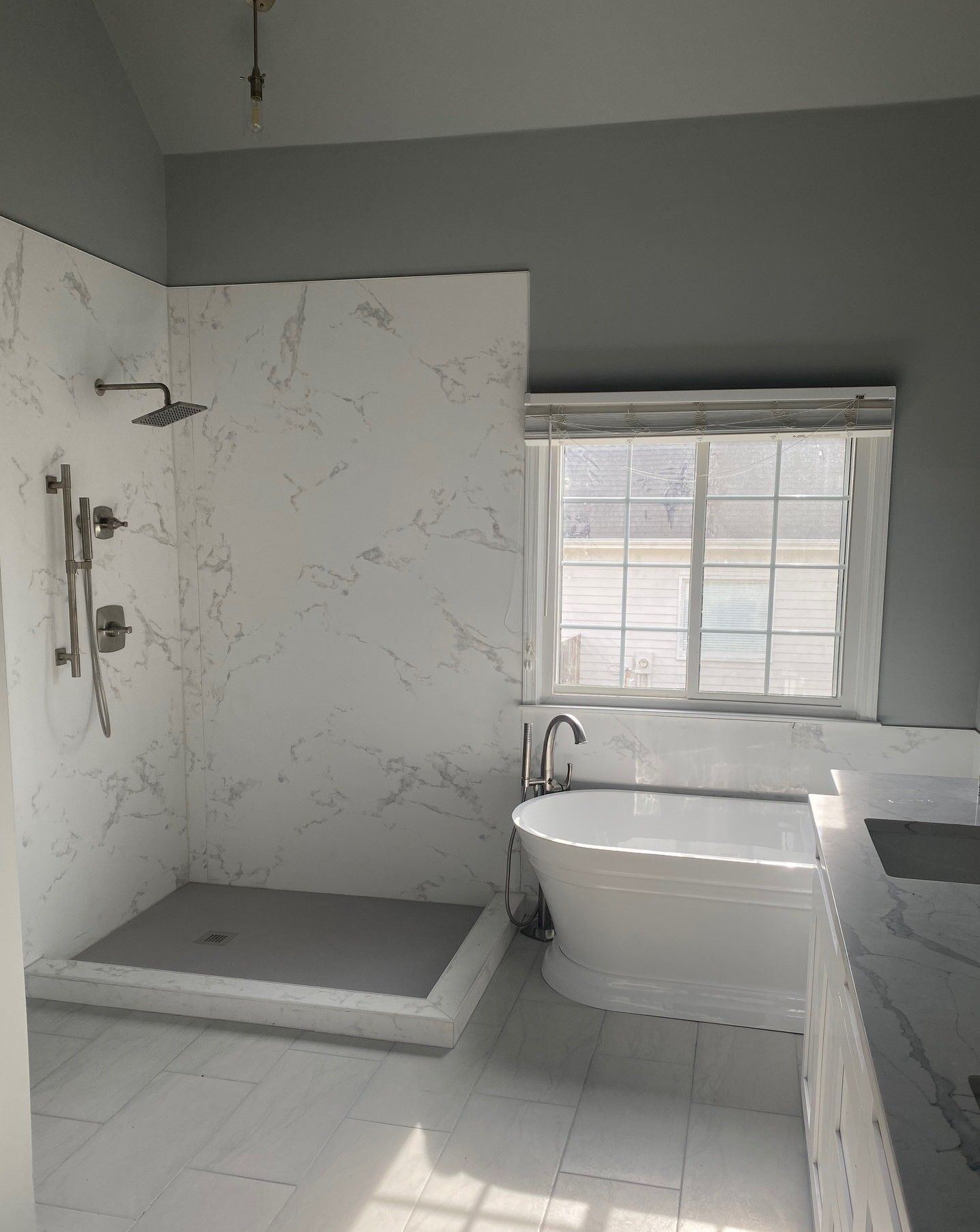 Modern bathroom with white marble-like tile. Shower, soaking tub, window, and grey walls.