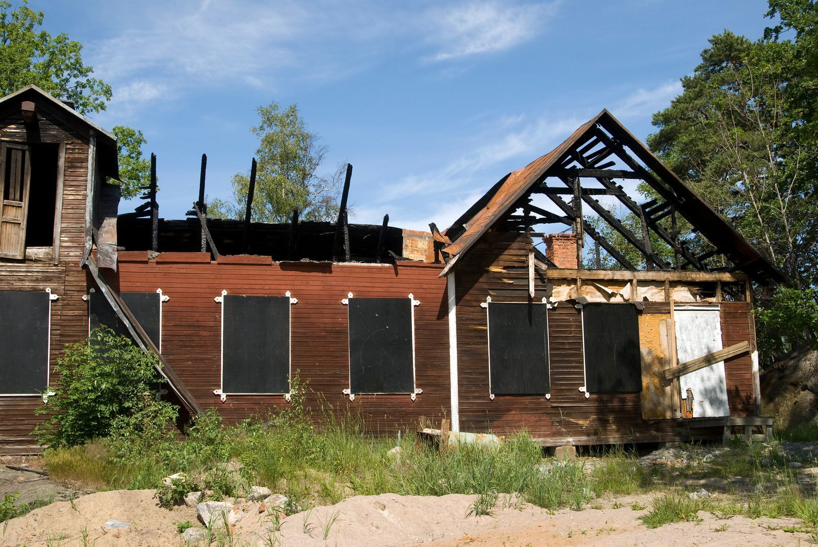 Burnt-out, red wooden house with boarded-up windows and exposed roof beams against a sunny, blue sky.