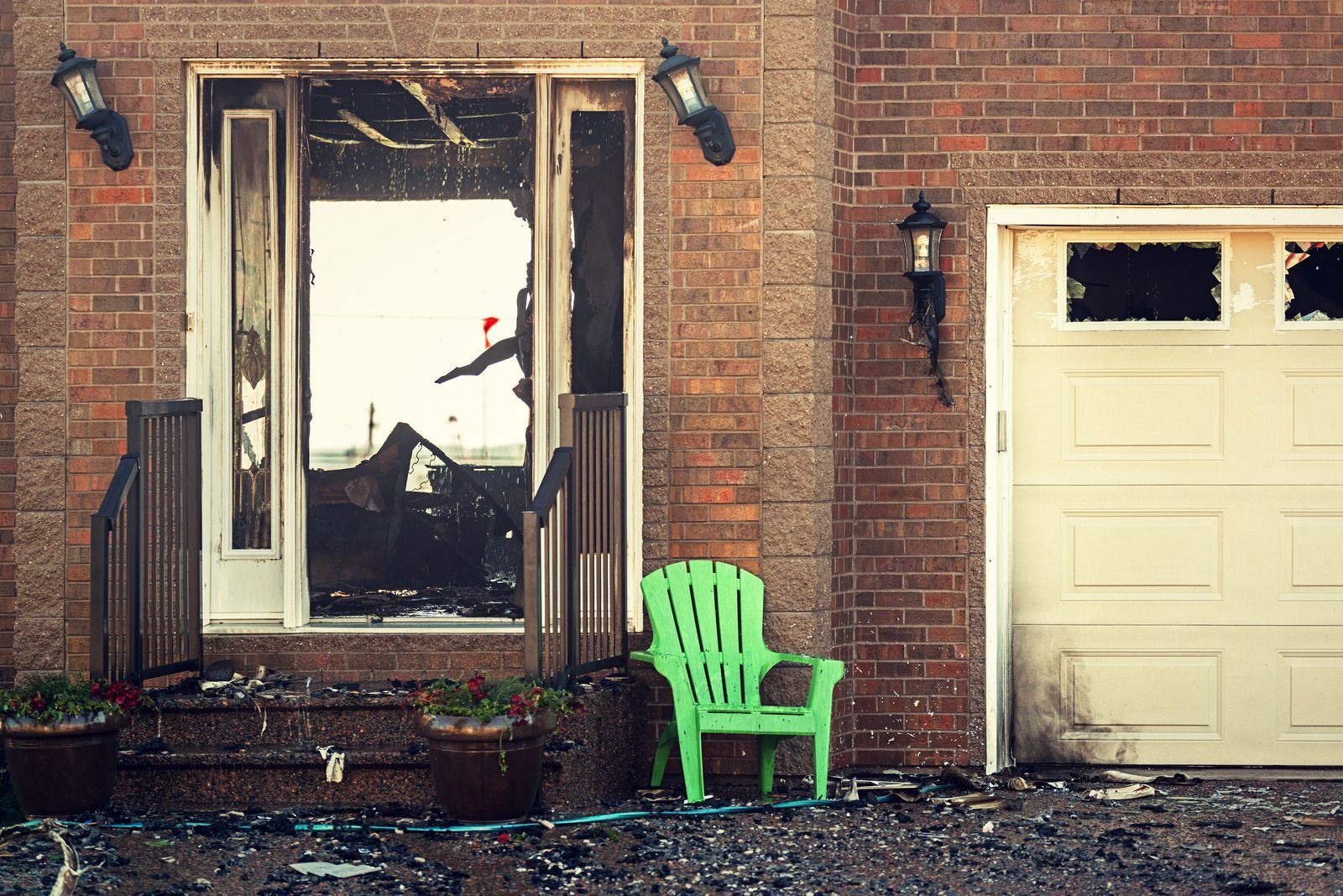 Exterior of a house with fire damage: charred front door, broken windows, and debris on the ground; a green chair sits nearby.