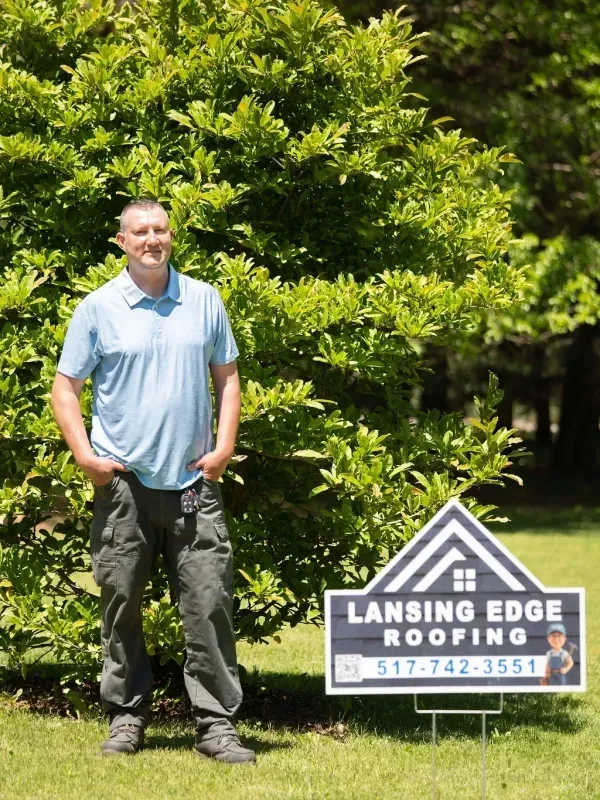 Man standing beside Lansing Edge Roofing yard sign in front of green trees