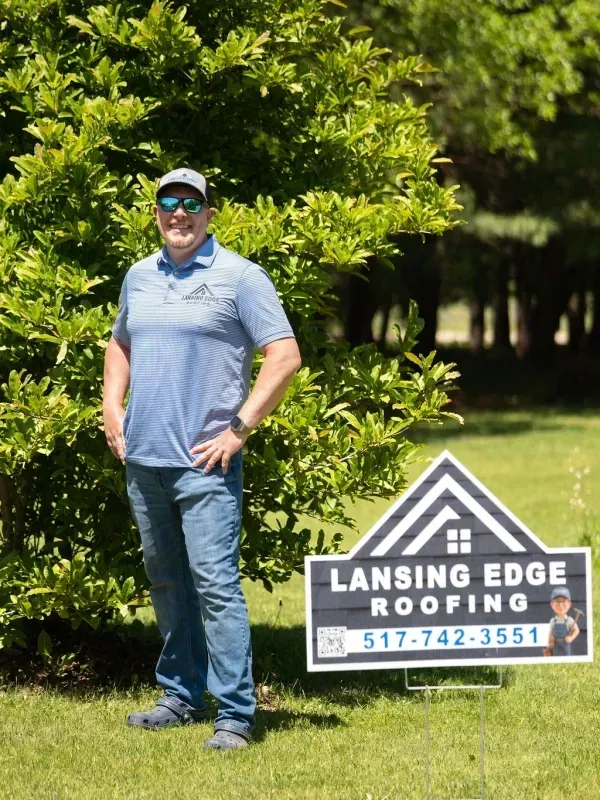 Man standing in a blue shirt beside a Lansing Edge Roofing sign on a sunny lawn